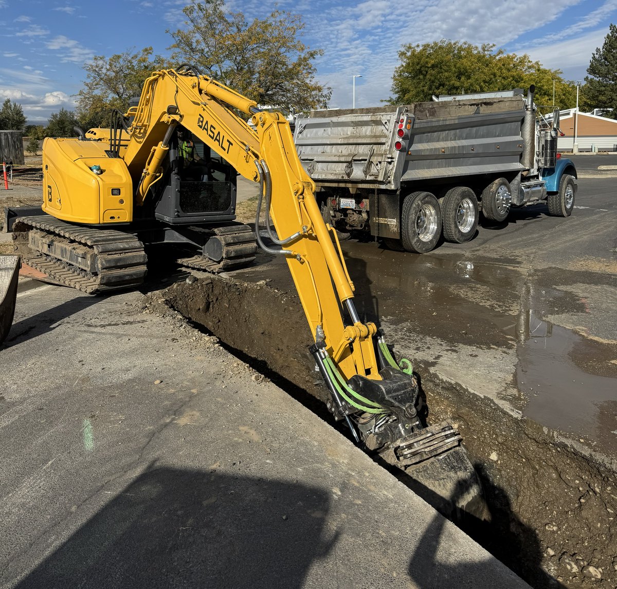 Basalt excavator with tiltrotator digging an open water main crossing trench with a dump truck alongside