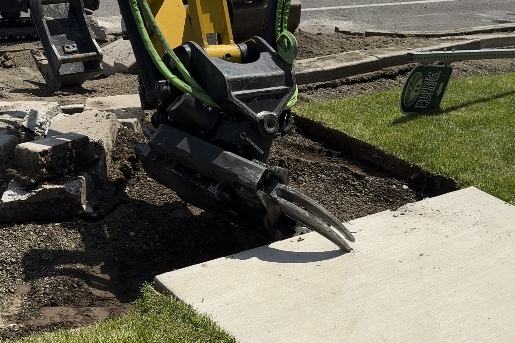 Tiltrotator grapple lifting a removed concrete panel beside a new sidewalk
