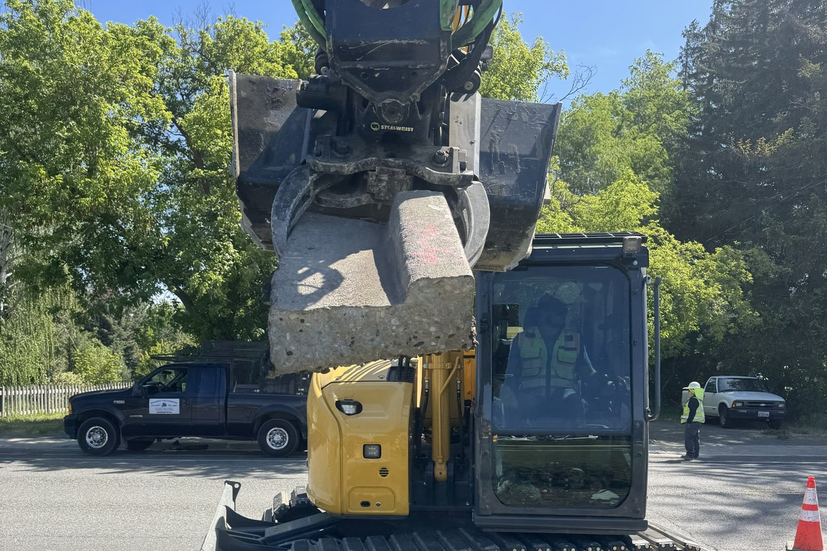 Tiltrotator grapple lifting a removed concrete curb panel under traffic control